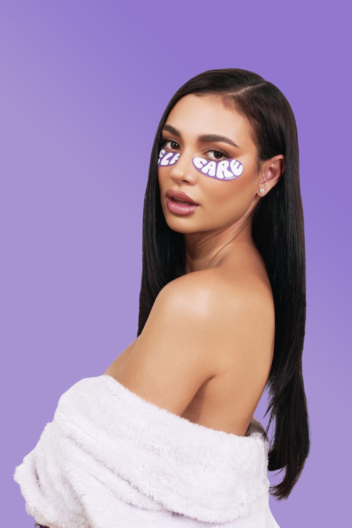 A woman looks over a bare shoulder at the camera, with a robe wrapped around her midsection. On her face is a pair of undereye masks. One says "Self" and the other says "Care."