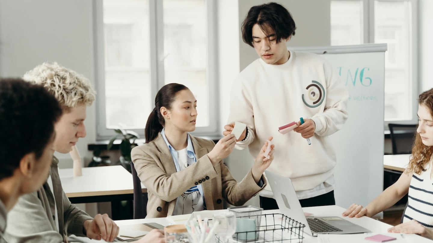 Fashion marketing professionals at work in an office, one stands around a table, the rest sit. The standing person hands another a sticky note. The room is well lit.