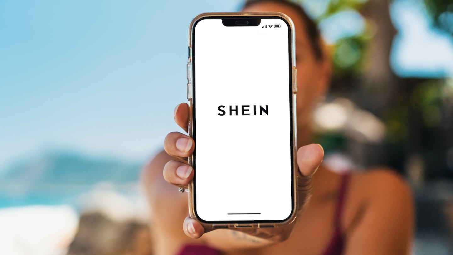 A woman on a beach holds up a phone with the Shein app open.