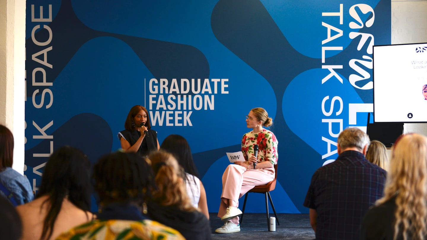 The image captures an indoor event at Graduate Fashion Week. The setting features a seated audience facing two speakers, likely engaged in a talk or presentation. One speaker is on stage, holding a microphone, while the backdrop displays “TALK SPACE” and “GRADUATE FASHION WEEK.” Attendees appear attentive, emphasising the educational and industry-focused context.